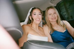 Radiant bride wearing lace veil sits with smiling woman in blue dress in car before wedding.