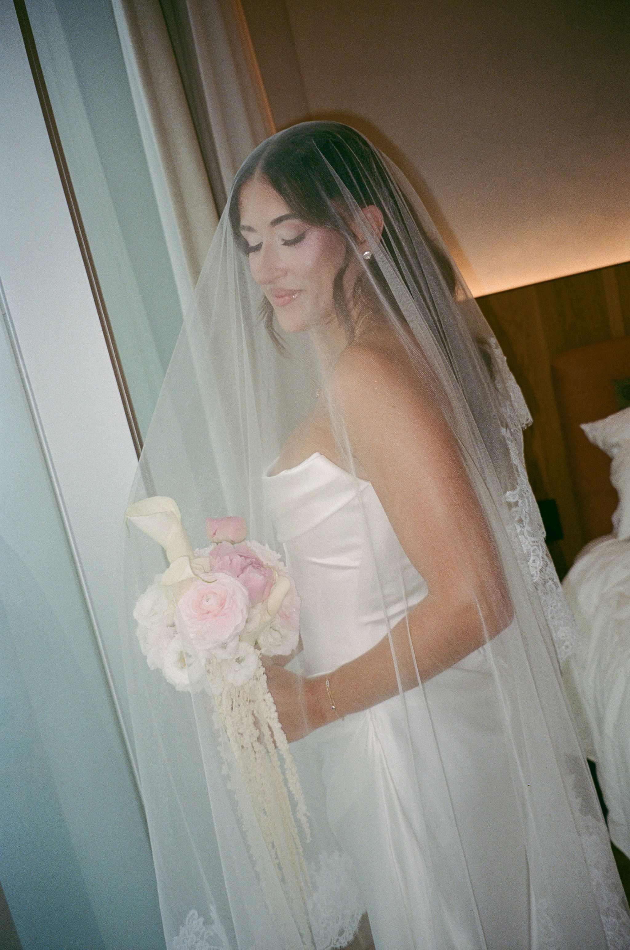 Elegant bride in strapless white gown holds pink and white bouquet with cascading details by window.