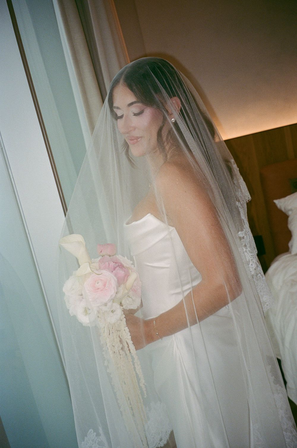 Elegant bride in strapless white gown holds pink and white bouquet with cascading details by window.
