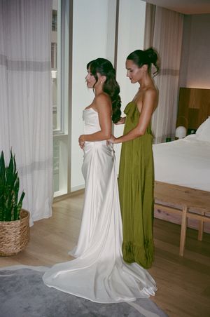 Bridesmaid in olive green dress fastens back of bride's white satin wedding gown in modern hotel room.