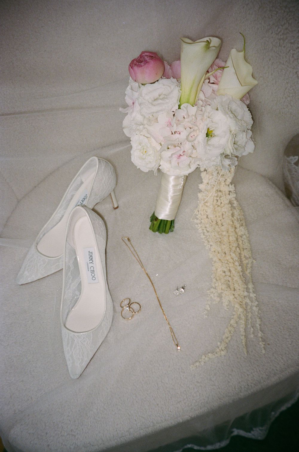 Flat lay of lace Jimmy Choo bridal heels, gold rings, necklace, and white pink flower bouquet.