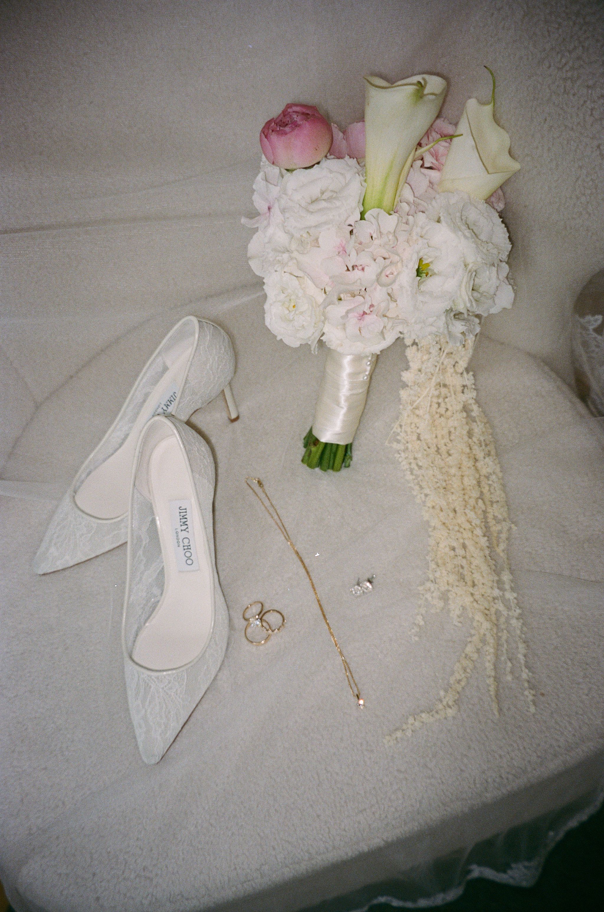 Flat lay of lace Jimmy Choo bridal heels, gold rings, necklace, and white pink flower bouquet.