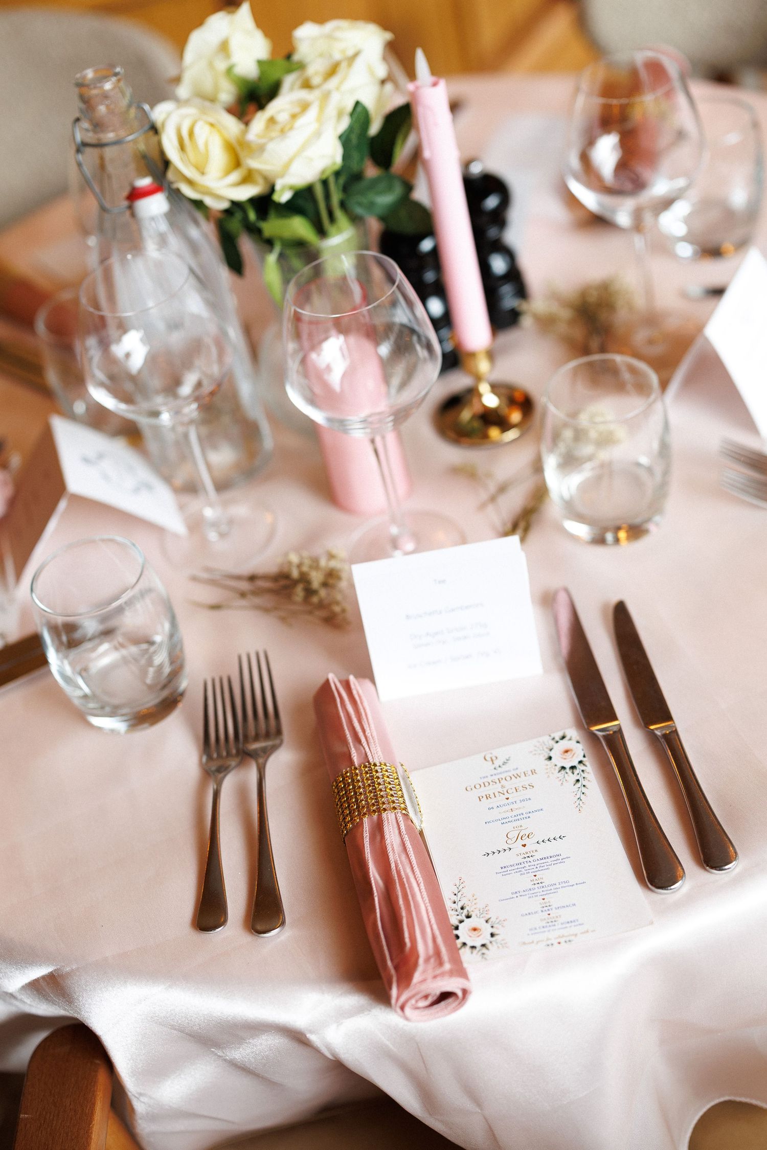 Elegant wedding table setting with pink napkin, gold cutlery, white roses, candles, and a menu card on blush linen.