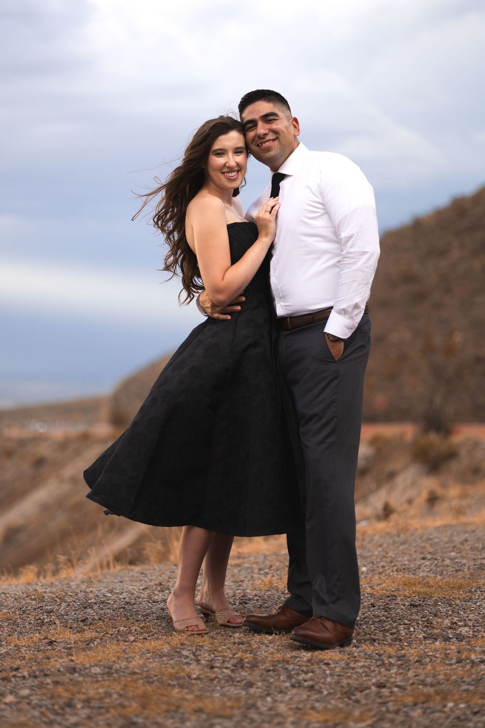 Couple posing outdoors in desert landscape, woman in black dress and man in white shirt with tie, smiling together.