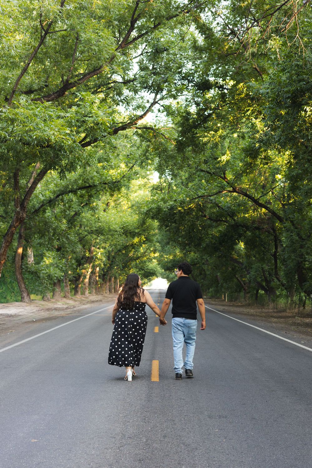 Couple holding hands walking down a tree-lined road, woman in polka dot dress and man in black shirt.