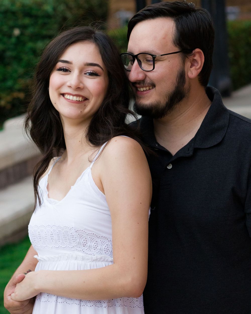 A smiling young couple poses together outdoors, woman in white dress and man in black polo shirt wearing glasses.