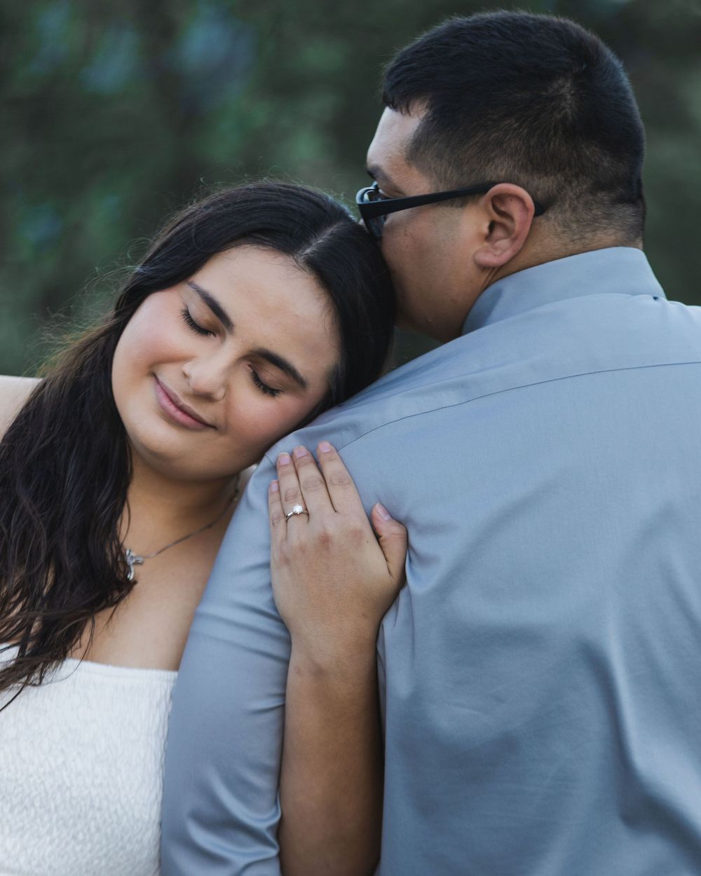 Couple sharing a tender embrace, woman smiling with engagement ring visible, man kissing her head outdoors.