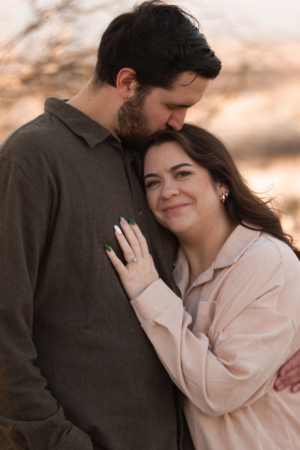 Couple embracing outdoors at golden hour, woman smiling in pink top with engagement ring, man kissing her head in olive shirt.