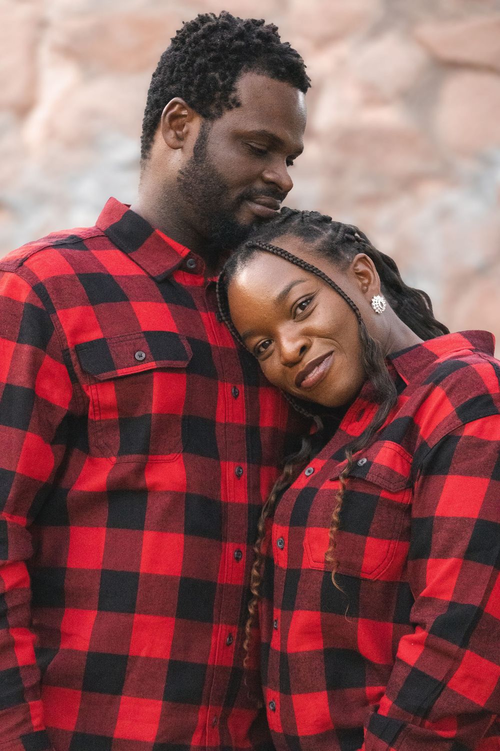 A loving Black couple in matching red plaid shirts embrace tenderly, the woman resting her head on the mans shoulder.