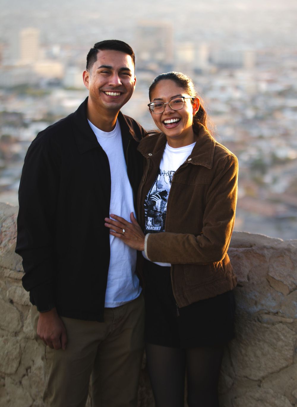 A smiling couple poses together on a scenic overlook with a city skyline in the background at golden hour.