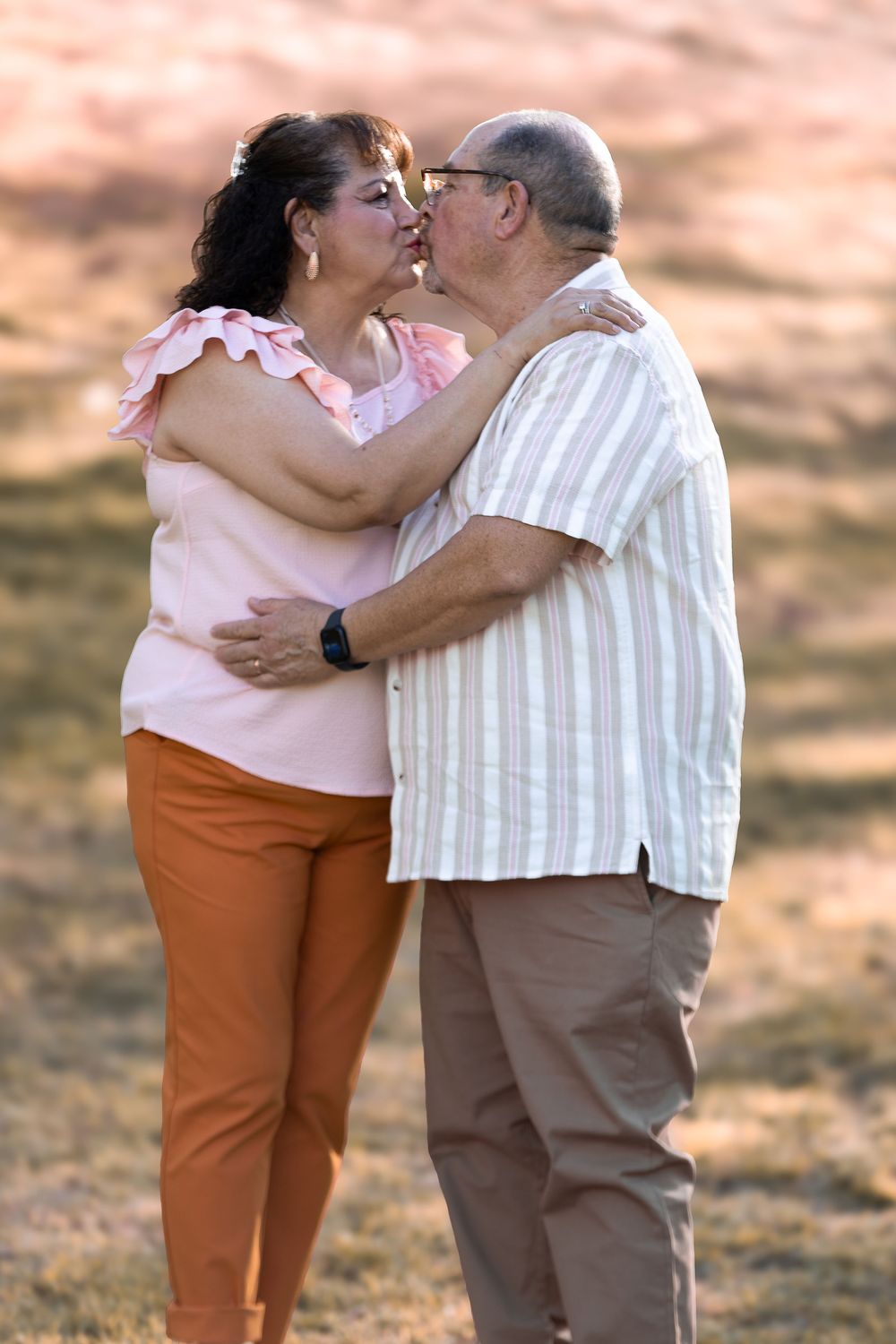 An older couple shares a romantic kiss while embracing outdoors in a park setting, woman in pink top and orange pants.