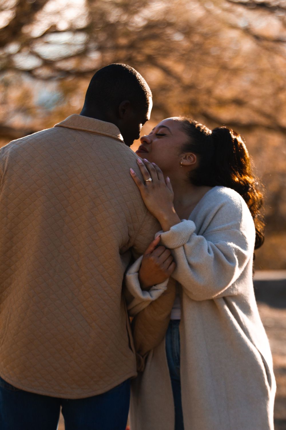 A couple shares a tender kiss outdoors in autumn, the woman wearing a ring and cozy coat, embraced in warm golden light.