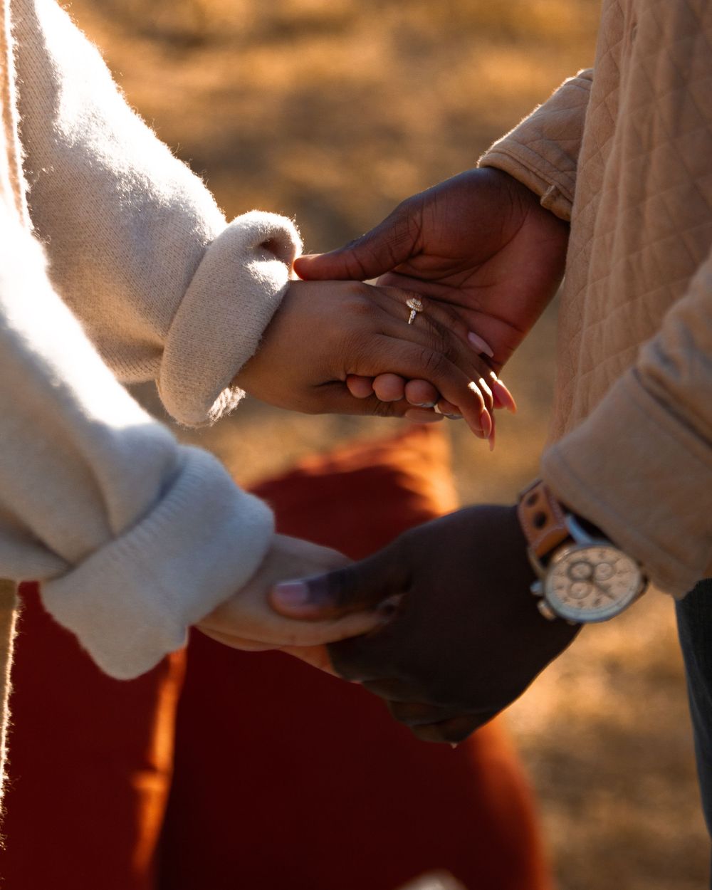 Two people holding hands outdoors in warm golden light, one wearing a watch and the other showing a ring on their finger.