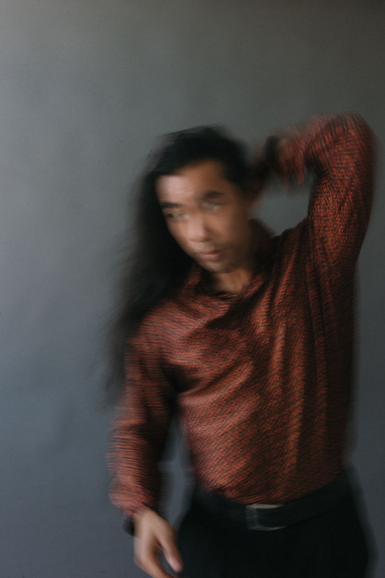 Blurred motion portrait of a long-haired Asian man in a red patterned shirt posing against a gray wall.