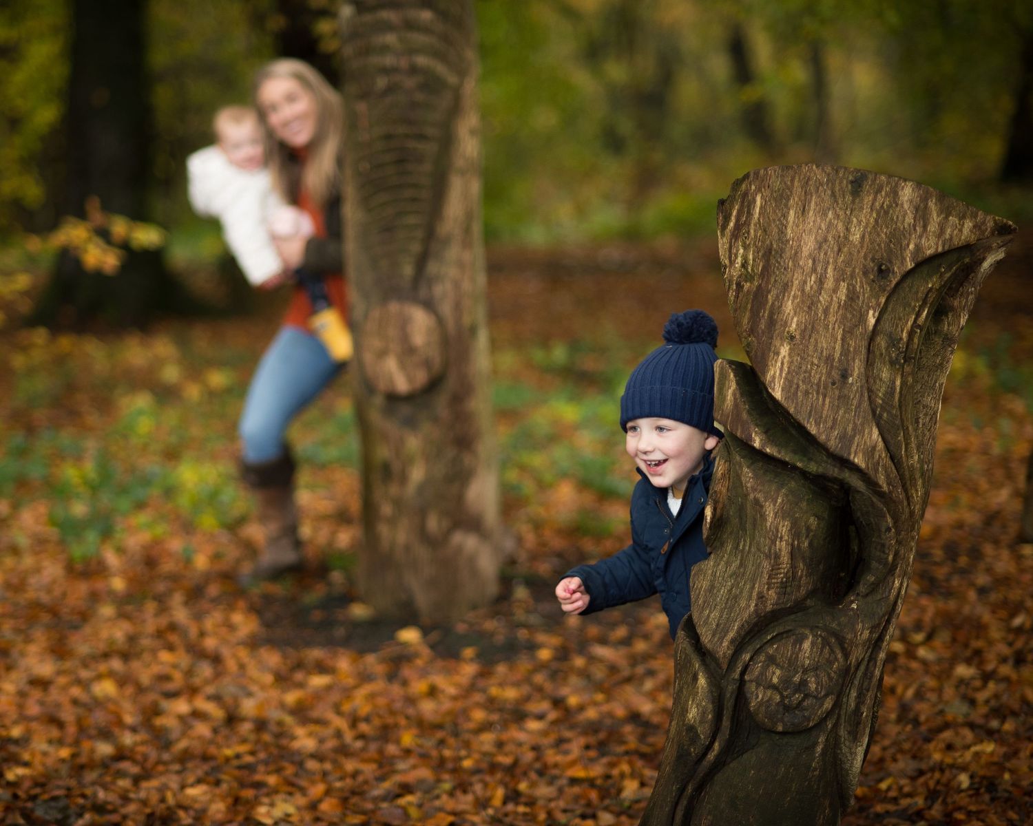 In an autumn forest, a smiling boy in a navy knit hat peeks from a carved tree stump while a woman holds a baby behind him.