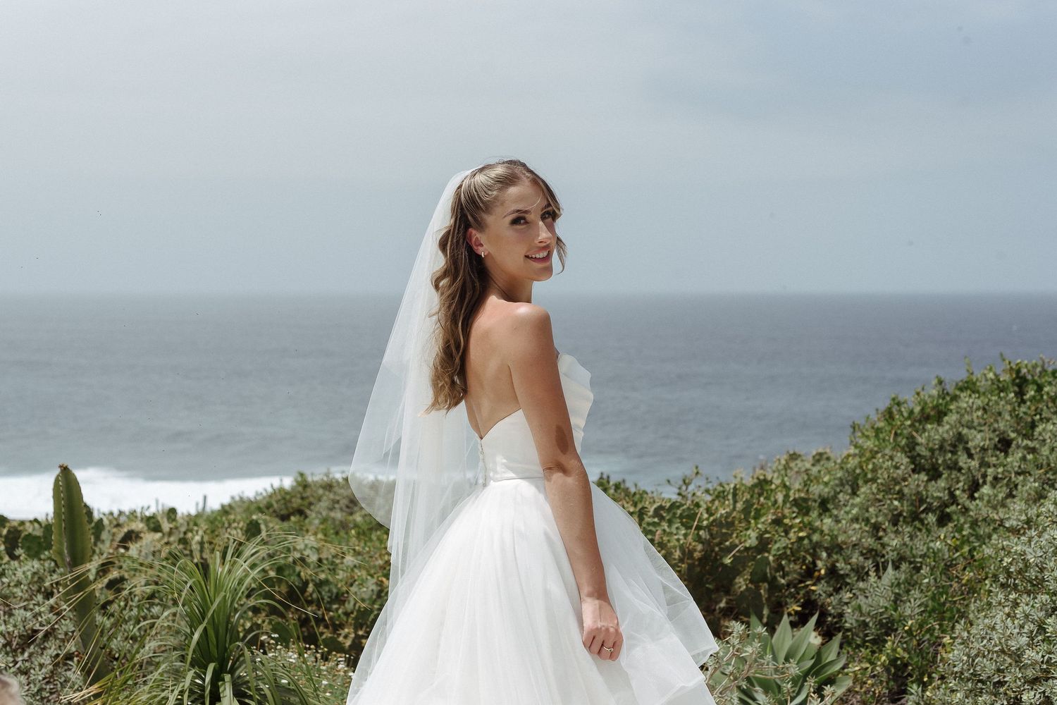 Bride in white gown and veil walks toward groom in beige suit on scenic coastal garden path overlooking the sea.