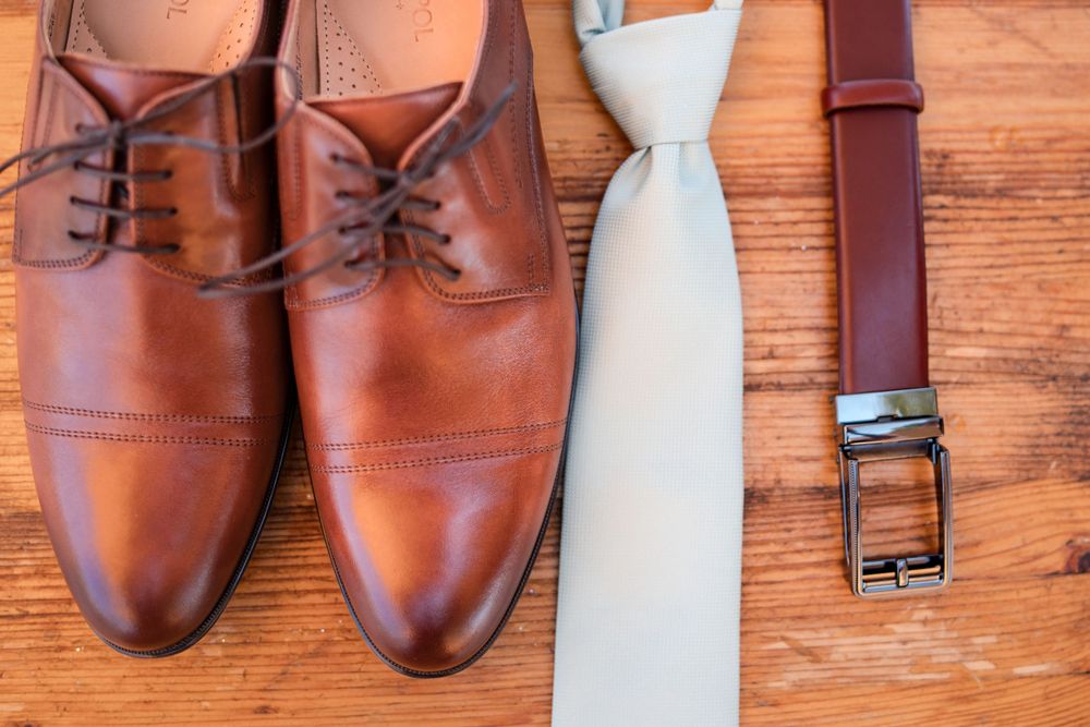Brown leather Oxford shoes, gray tie, and brown belt arranged on wooden surface for groom wedding attire.