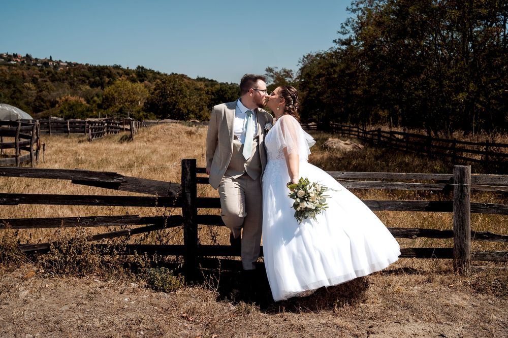 Bride and groom kissing near a rustic wooden fence on a sunny day at a countryside wedding venue.