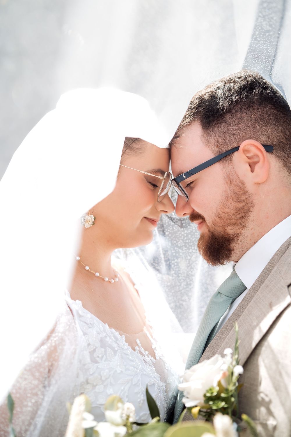 A bride and groom touch foreheads tenderly under a flowing white veil, sharing an intimate moment on their wedding day.