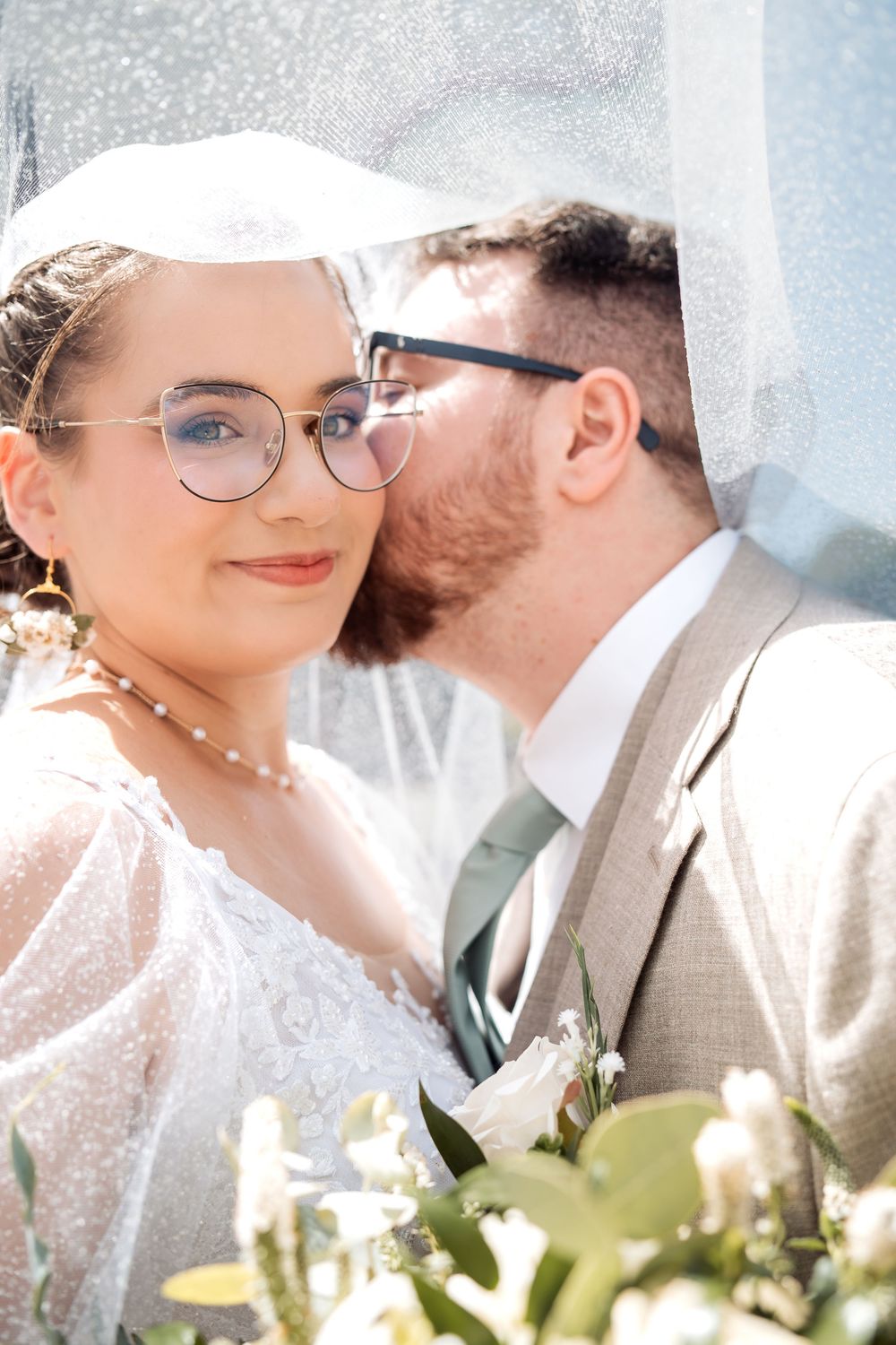 Bride and groom share a tender moment under a veil, both wearing glasses, with white floral bouquet in foreground.