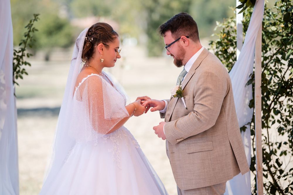 Bride and groom exchanging rings during outdoor wedding ceremony, bride in white gown with veil, groom in tan suit.