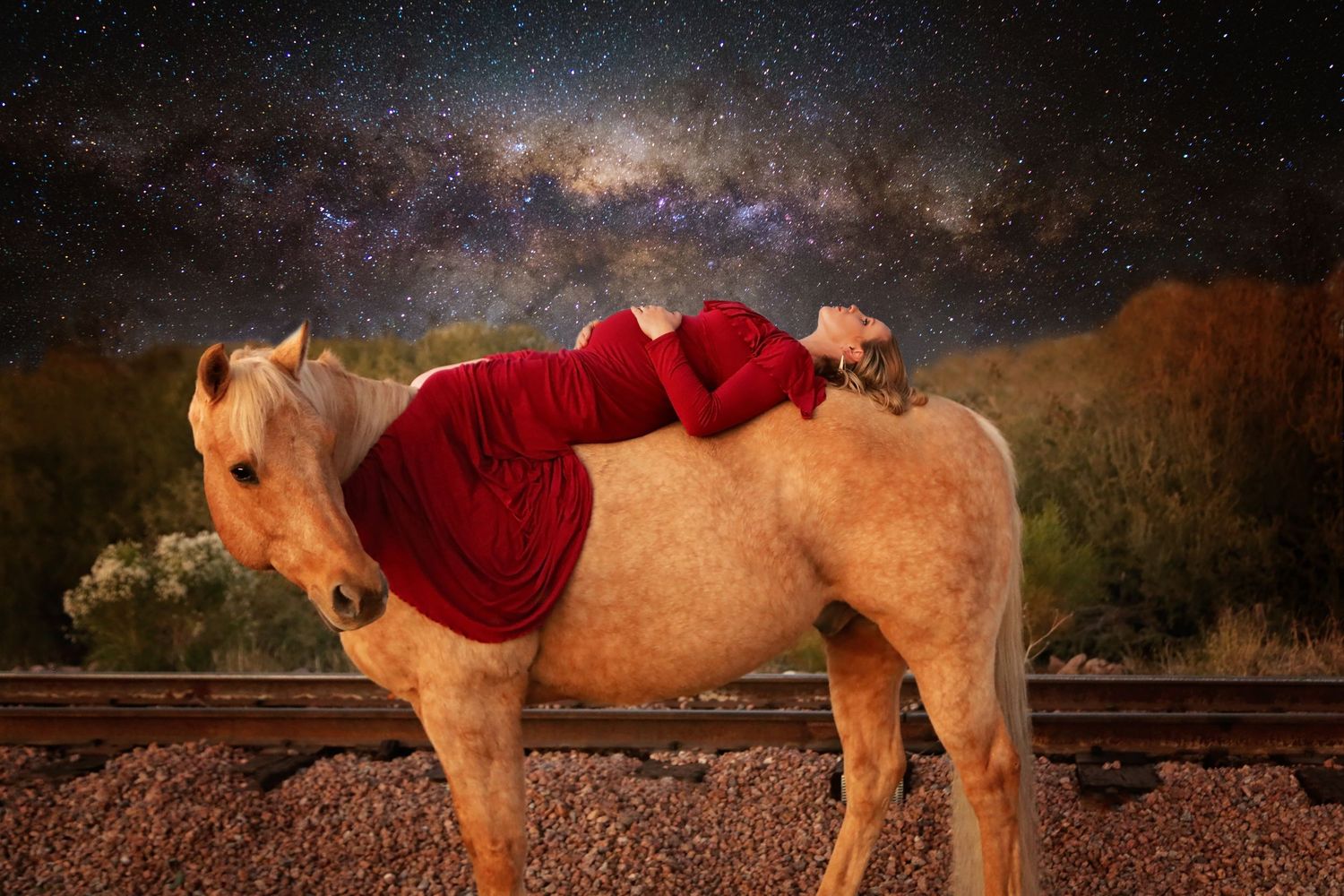 Woman in a flowing red dress rests on a palomino horse by railroad tracks beneath a starry Milky Way night sky.