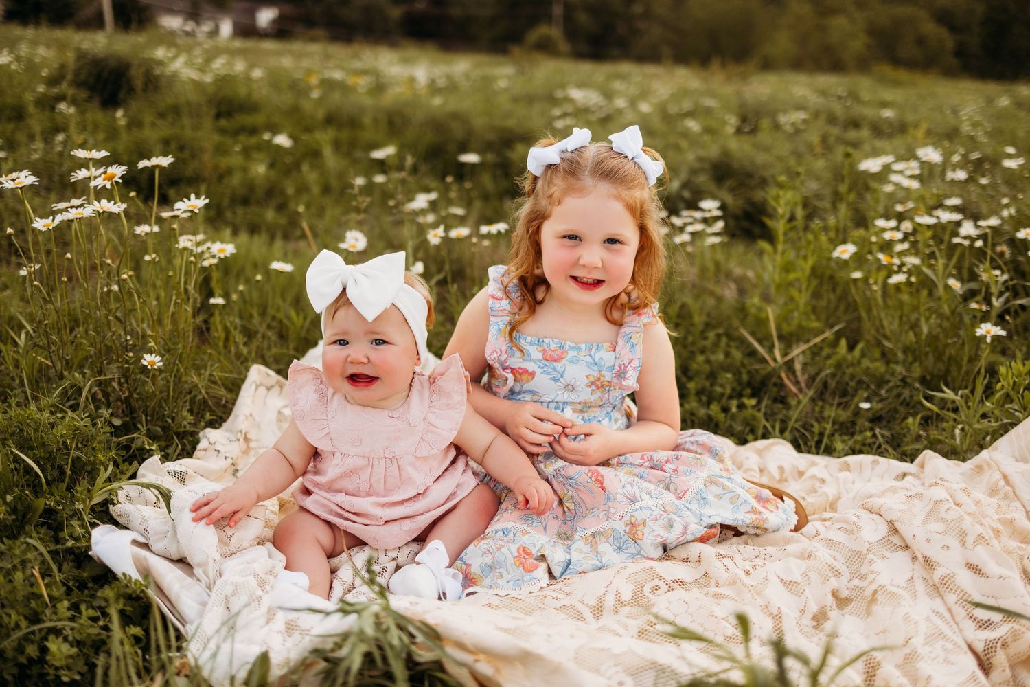 Two young sisters in floral dresses sit on a blanket in a daisy field, wearing white hair bows.