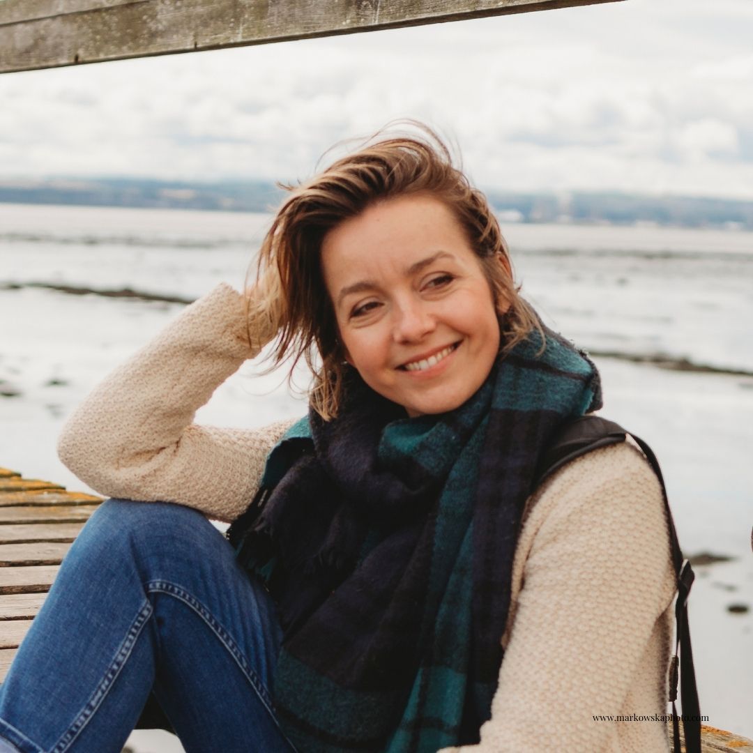 Person in cream sweater and plaid scarf sitting outdoors by the ocean.