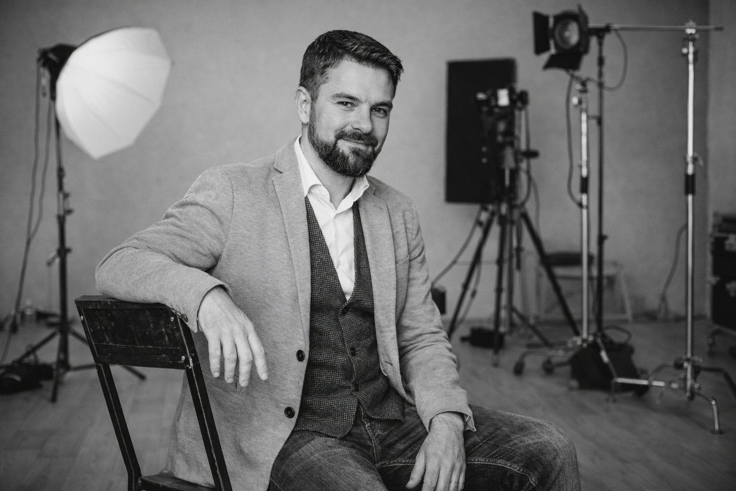 Smiling bearded man in suit jacket seated in photography studio with professional lighting equipment.