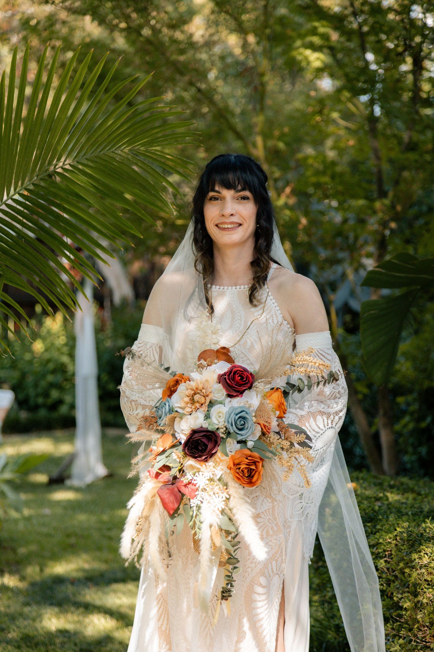 Smiling bride in boho off-shoulder gown holds colorful bouquet with roses outdoors among lush greenery.