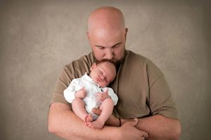 Bald father kissing sleeping newborn baby dressed in white, cradled in his arms against a gray background.