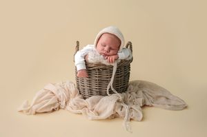 Newborn baby sleeping peacefully in a rustic wicker basket with soft cream fabric on a beige background.