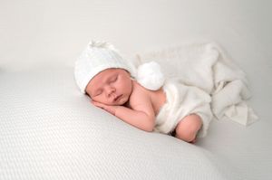 Peaceful newborn baby sleeping in a white knit hat and blanket on a soft white surface.