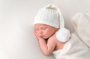 Peaceful newborn baby sleeping in white knit hat with pompom, wrapped in soft white blanket.