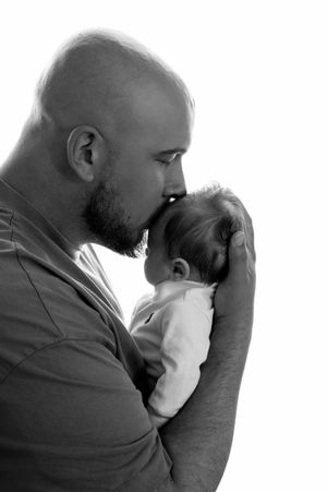 Bald father with beard tenderly kissing newborn baby's head in black and white portrait photo.