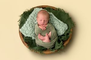 Newborn baby sleeping peacefully wrapped in green cloth inside a wooden bowl with leafy green decor.