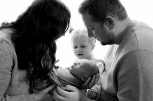Black and white family portrait of parents holding newborn baby with toddler sibling smiling.