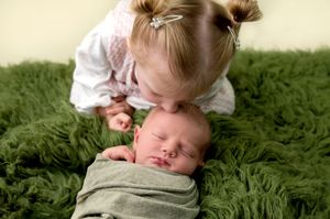 Toddler girl with pigtails kissing sleeping newborn baby sibling wrapped in green swaddle on fluffy green blanket.
