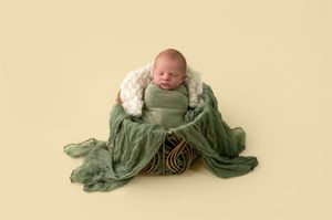 Newborn baby wrapped in green fabric sleeping in a wicker basket with white fur, on a yellow background.