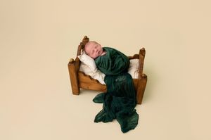 Newborn baby wrapped in green velvet swaddle sleeping on tiny wooden bed with white pillow.