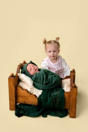 Toddler girl with pigtails stands behind a wooden doll bed holding a swaddled newborn baby wrapped in green fabric.