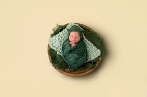 Newborn baby wrapped in green swaddle with matching hat, nestled in a wooden bowl with green leaves on cream background.