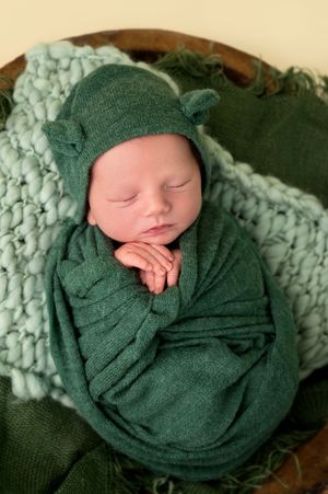 Sleeping newborn baby swaddled in green wrap with elf ear hat posed in wooden bowl on teal knit blanket.