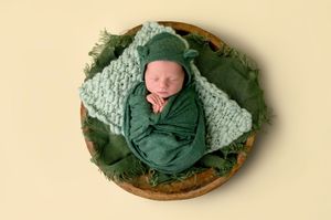 Newborn baby swaddled in green wrap with bear hat, sleeping peacefully in a wooden bowl with green layers.