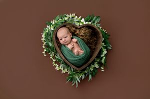 Newborn baby wrapped in green swaddled in a heart-shaped wooden bowl with floral greenery on brown background.