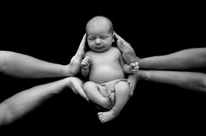 Black and white photo of a newborn baby cradled by multiple adult hands against a dark background.