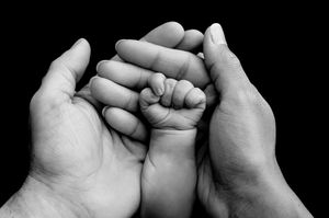 Black and white photo of a baby's tiny fist resting in the open palms of two adult hands.