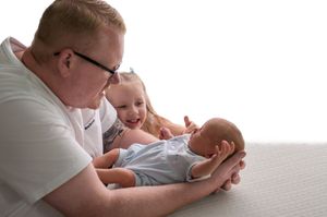 Adult holding newborn baby while young child smiles nearby on white surface.
