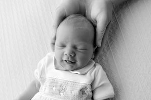 Black and white photo of a sleeping newborn baby smiling, with a parent's hand gently resting on its head.