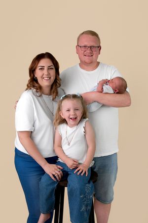 Family portrait with parents and two children, including a newborn baby, wearing white tops and jeans on beige background.